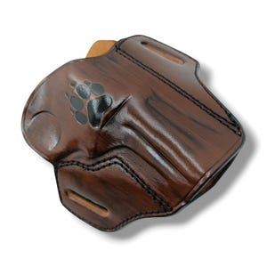 May include: A brown leather holster with a paw print design. The holster has a glossy finish and black stitching. It features a belt loop and a molded shape for a firearm.