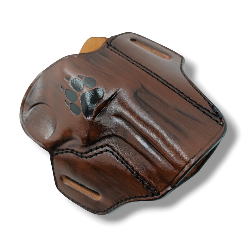 May include: A brown leather holster with a paw print design. The holster has a glossy finish and black stitching. It features a belt loop and a molded shape for a firearm.