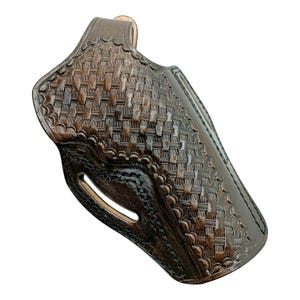 Op de afbeelding: Een bruine leren holster met een mandweefpatroon. De holster heeft een gebogen vorm met een riemlus en een uitsparing voor de trekkerbeugel van het vuurwapen. Het leer heeft een glanzende afwerking.