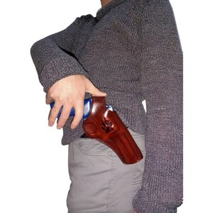 Peut inclure: Un &eacute;tui en cuir marron avec un motif de patte. L'&eacute;tui est con&ccedil;u pour contenir une arme de poing bleue. L'image montre l'&eacute;tui port&eacute; &agrave; la hanche.