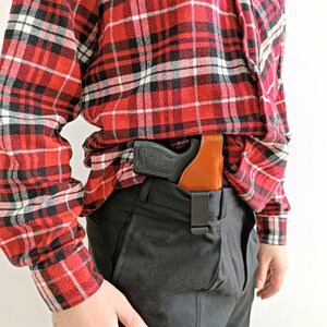 Op de afbeelding: Een zwart en bruin holster met een zwart pistool is vastgeklikt aan de tailleband van een zwarte broek. De persoon draagt een rood, zwart en wit geruit flanellen overhemd.