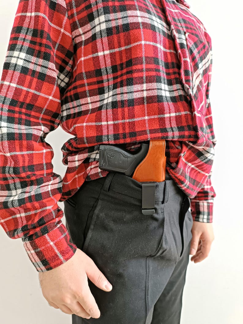 Op de afbeelding: Een zwart en bruin holster met een zwart pistool is vastgeklikt aan de tailleband van een zwarte broek. De persoon draagt een rood, zwart en wit geruit flanellen overhemd.