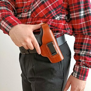 Op de afbeelding: Een bruin leren holster met een zwart pistool, vastgeklikt aan een zwarte riem. Het holster is zichtbaar tegen een rood-zwart geruit overhemd en een zwarte broek.