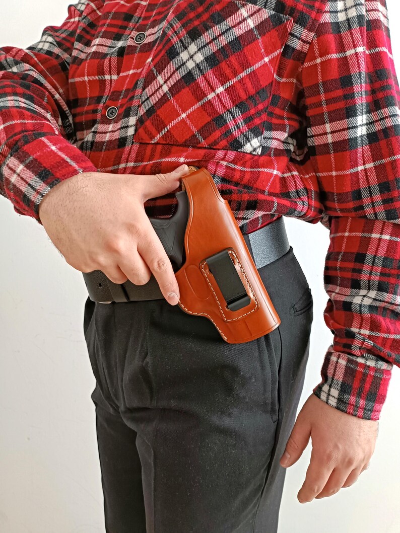 Op de afbeelding: Een bruin leren holster met een zwart pistool, vastgeklikt aan een zwarte riem. Het holster is zichtbaar tegen een rood-zwart geruit overhemd en een zwarte broek.