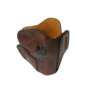May include: A brown leather mask with a paw print design. The mask has a dark brown exterior with a lighter brown interior. The paw print is black. The mask has a curved shape and stitched edges.