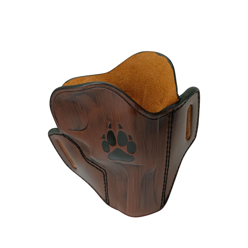 May include: A brown leather mask with a paw print design. The mask has a dark brown exterior with a lighter brown interior. The paw print is black. The mask has a curved shape and stitched edges.