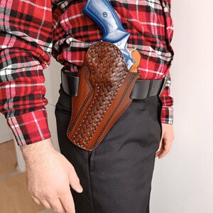 Op de afbeelding: Een bruin leren holster met een geweven ontwerp, met een blauw en zilveren revolver. Het holster is bevestigd aan een zwarte riem die over een zwarte broek wordt gedragen. De persoon draagt een rood-zwart geruit overhemd.