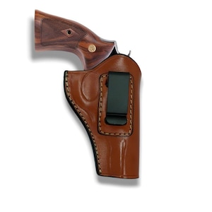 Op de afbeelding: Een bruin leren holster met een zwarte clip, met een revolver met een houten handvat. De handgreep van de revolver heeft een donker houtnerfpatroon en een gouden embleem. De holster is gestikt met een lichtere bruine draad.