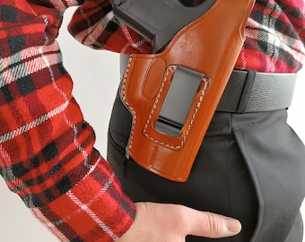 Op de afbeelding: Een bruin leren holster met een zwarte clip is bevestigd aan een zwarte riem. Een zwart pistool is gedeeltelijk zichtbaar in het holster. De persoon draagt een rood-zwart geruit overhemd en een zwarte broek.