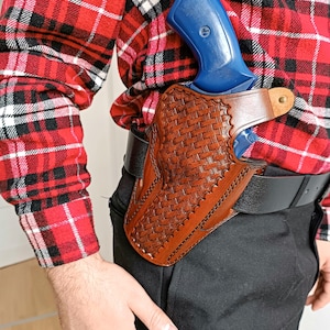 Könnte beinhalten: Eine braune Lederholster mit einem geflochtenen Korbdesign, die eine blaue Schusswaffe hält. Die Holster ist an einem schwarzen Gürtel befestigt, der mit einer schwarzen Hose und einem rot-schwarz karierten Hemd getragen wird.