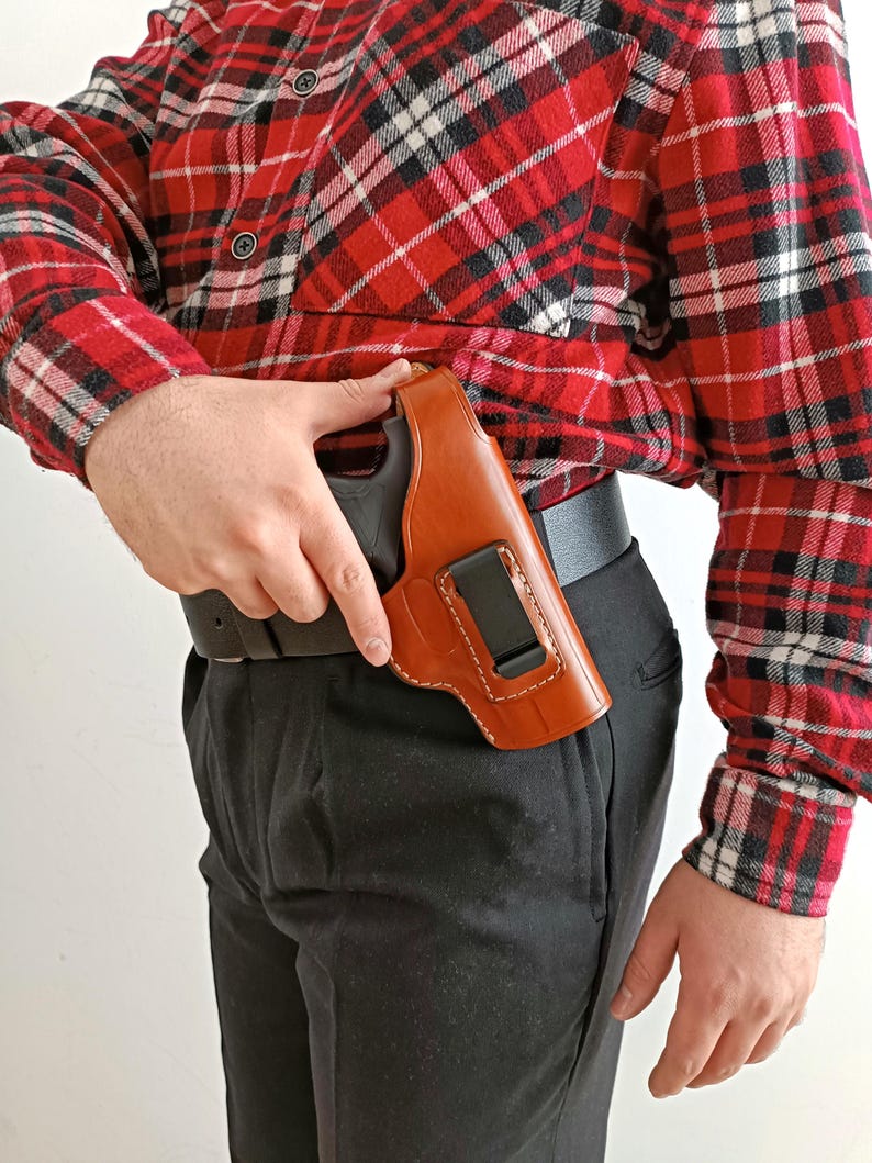 K&ouml;nnte beinhalten: Ein braunes Lederholster mit einer schwarzen Schusswaffe, die an einem schwarzen G&uuml;rtel befestigt ist. Das Holster ist vor einem rot-schwarz karierten Hemd und einer schwarzen Hose zu sehen.
