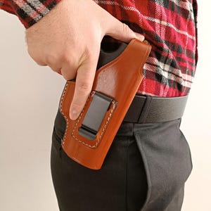 Op de afbeelding: Een bruin leren holster met witte stiksels en een zwarte metalen clip is bevestigd aan een zwarte riem. De holster wordt vastgehouden door een persoon die een rood-zwart geruit overhemd en een zwarte broek draagt.