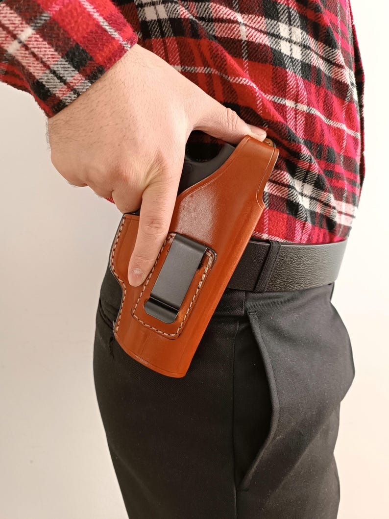 Op de afbeelding: Een bruin leren holster met witte stiksels en een zwarte metalen clip is bevestigd aan een zwarte riem. De holster wordt vastgehouden door een persoon die een rood-zwart geruit overhemd en een zwarte broek draagt.