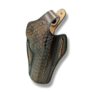 Op de afbeelding: Een donkerbruin leren holster met een mandweefpatroon. Het holster is gevormd voor een vuurwapen, met een riemlus en uitsparingen. Het leer heeft een getextureerde uitstraling.