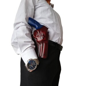 Peut inclure: Un &eacute;tui en cuir bordeaux avec un motif de patte, contenant un revolver &agrave; poign&eacute;e bleue. L'&eacute;tui est port&eacute; sur une ceinture noire, et la personne porte une chemise blanche et un pantalon noir. Une montre argent&eacute;e est visible.