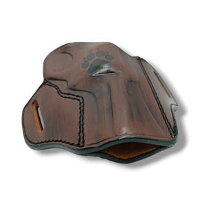 May include: A brown leather holster with a paw print design. The holster has black stitching and a smooth, polished finish. The interior is a lighter brown color. The holster is designed to hold a firearm.