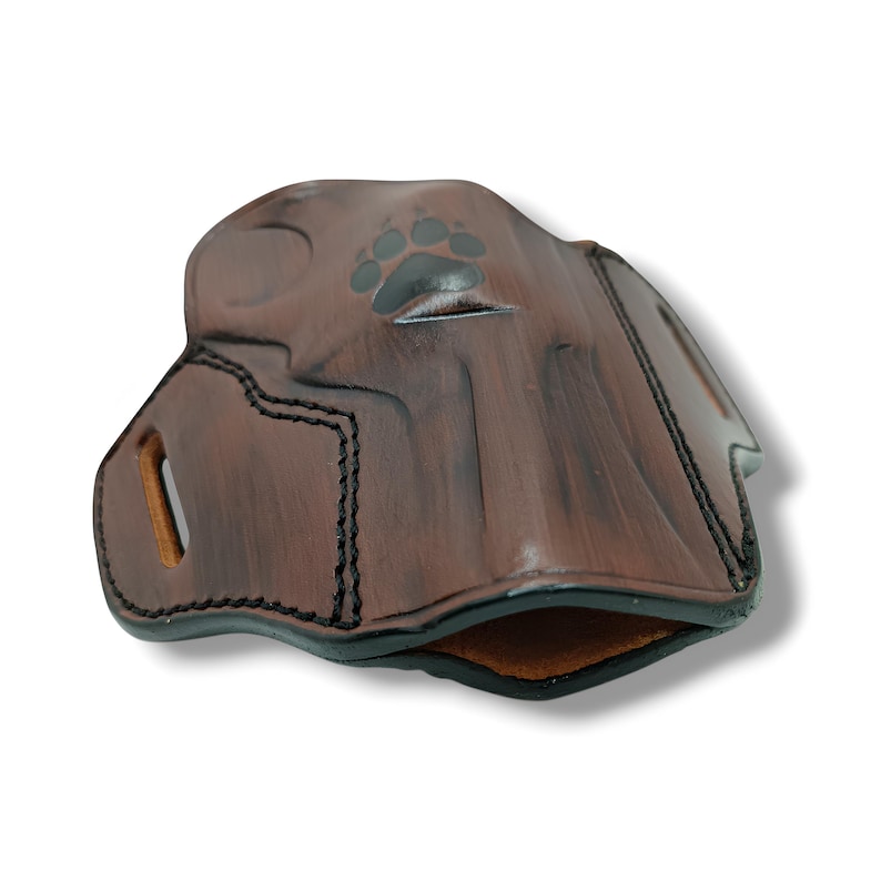 May include: A brown leather holster with a paw print design. The holster has black stitching and a smooth, polished finish. The interior is a lighter brown color. The holster is designed to hold a firearm.