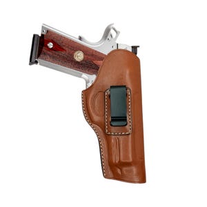 Op de afbeelding: Een zilveren pistool met een houten handvat is gedeeltelijk in een bruine leren holster gestoken. De holster heeft een zwarte clip en witte stiksels. Het pistool heeft een zwart magazijn.