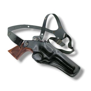 Peut inclure: Un holster d'épaule en cuir noir avec un revolver à manche marron. L'étui est moulé pour s'adapter à l'arme, et les sangles sont réglables. Le revolver est partiellement visible, montrant sa poignée en bois.