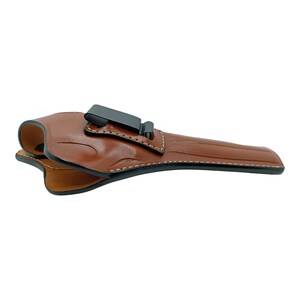 Op de afbeelding: Een bruin leren holster met een zwarte clip. De holster is ontworpen om een ​​handvuurwapen te bevatten. De randen zijn gestikt met witte draad. Het leer heeft een gladde, gepolijste afwerking.