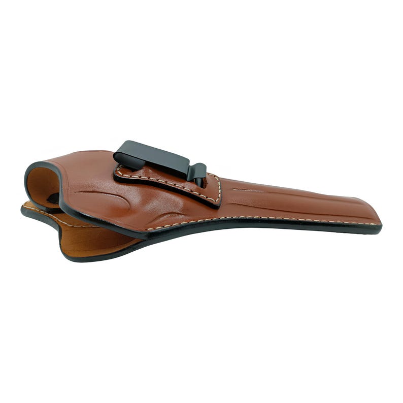 Op de afbeelding: Een bruin leren holster met een zwarte clip. De holster is ontworpen om een ​​handvuurwapen te bevatten. De randen zijn gestikt met witte draad. Het leer heeft een gladde, gepolijste afwerking.