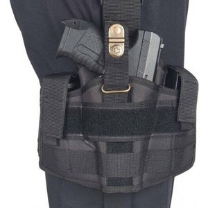 May include: Black tactical thigh holster with a handgun secured inside. The holster features multiple straps, a buckle, and a hook-and-loop closure. The holster is designed to be worn on the thigh and is made of durable, heavy-duty material.