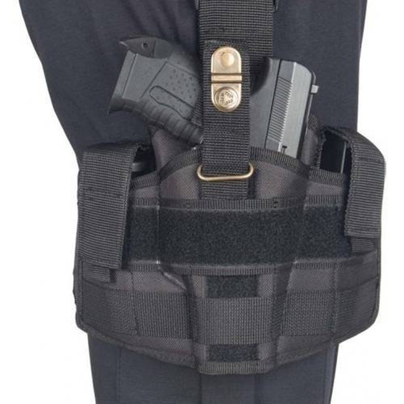 May include: Black tactical thigh holster with a handgun secured inside. The holster features multiple straps, a buckle, and a hook-and-loop closure. The holster is designed to be worn on the thigh and is made of durable, heavy-duty material.