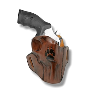 May include: A brown leather holster with a dark wood grain finish and a black paw print cutout design. The holster features black stitching along the edges and belt slots on both sides, containing a silver revolver with a black textured grip.