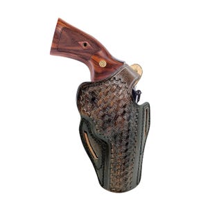 May include: A brown leather holster with a woven basketweave pattern, holding a revolver. The revolver has a wooden grip and a gold-coloured emblem. The holster is designed to be worn on the hip.