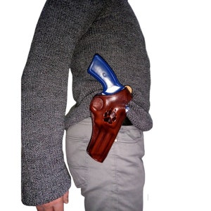 Peut inclure: Un &eacute;tui en cuir marron avec un motif de patte, contenant un revolver bleu et argent&eacute;. L'&eacute;tui est port&eacute; &agrave; la hanche, visible contre un pull et un pantalon gris.