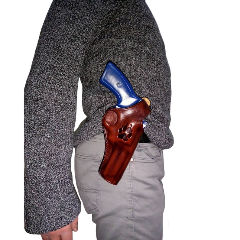 Peut inclure: Un &eacute;tui en cuir marron avec un motif de patte, contenant un revolver bleu et argent&eacute;. L'&eacute;tui est port&eacute; &agrave; la hanche, visible contre un pull et un pantalon gris.