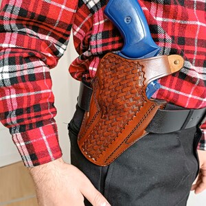 Op de afbeelding: Een bruine leren holster met een geweven mandontwerp, met een blauw pistool. De holster is bevestigd aan een zwarte riem en de persoon draagt een rood-zwart geruit overhemd.