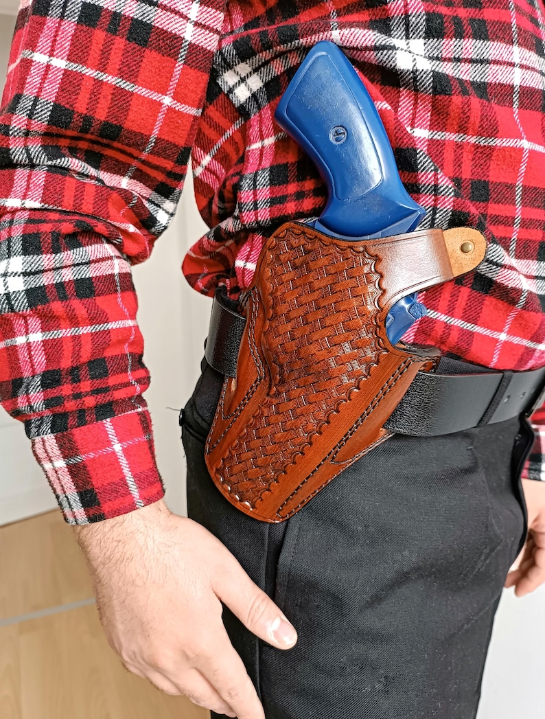 May include: A brown leather holster with a woven pattern, holding a blue handgun. The holster is attached to a black belt. The person is wearing a red and black plaid shirt and black pants.