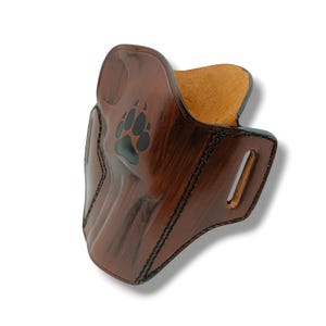 May include: A brown leather holster with a black bear paw print. The holster has a textured finish and black stitching along the edges. The interior is a lighter tan color, and there is a belt loop on the side.