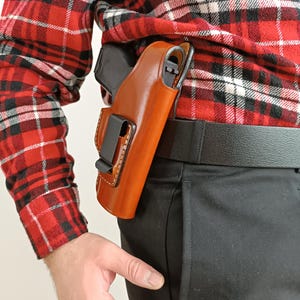 Op de afbeelding: Een bruin leren holster met een zwarte clip is bevestigd aan een zwarte riem. Een zwart pistool is gedeeltelijk zichtbaar. De persoon draagt een rood-zwart geruit overhemd en een zwarte broek. Het holster wordt aan de rechterkant gedragen.