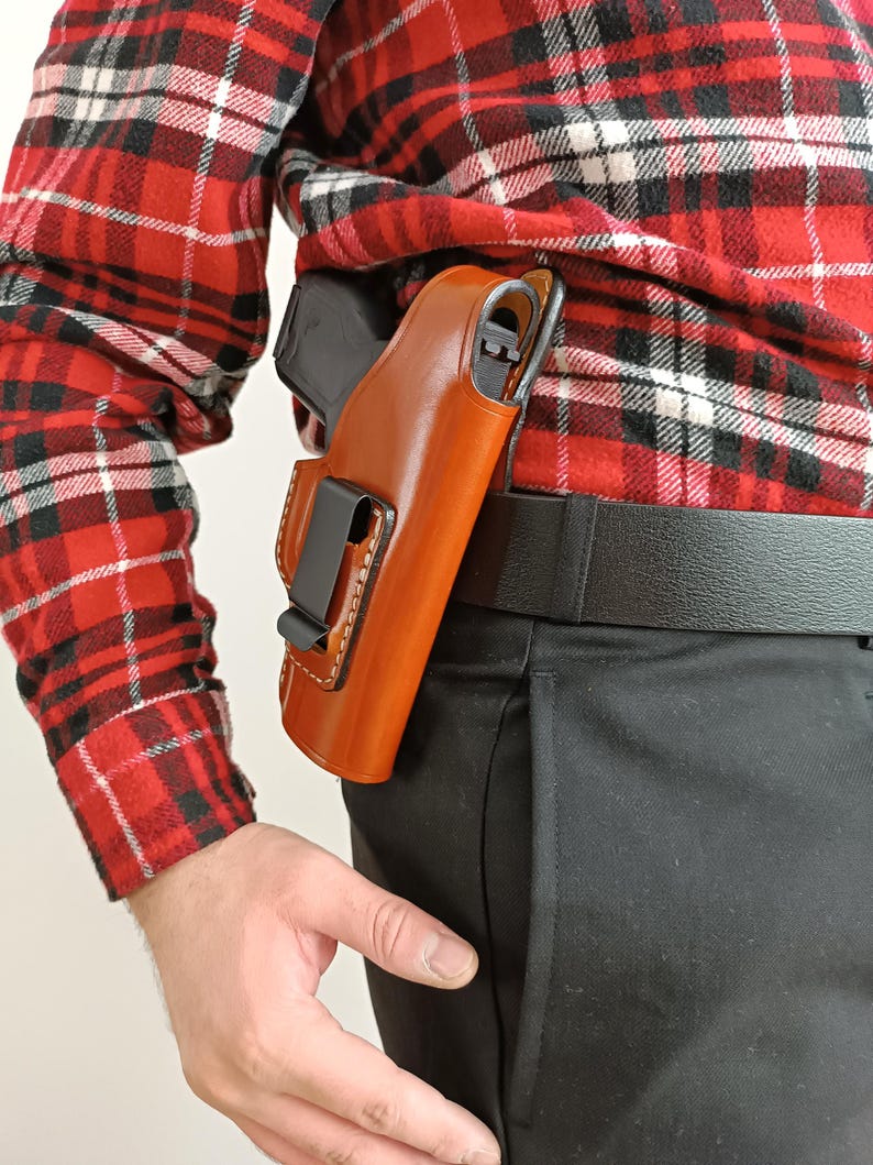 Op de afbeelding: Een bruin leren holster met een zwarte clip is bevestigd aan een zwarte riem. Een zwart pistool is gedeeltelijk zichtbaar. De persoon draagt een rood-zwart geruit overhemd en een zwarte broek. Het holster wordt aan de rechterkant gedragen.
