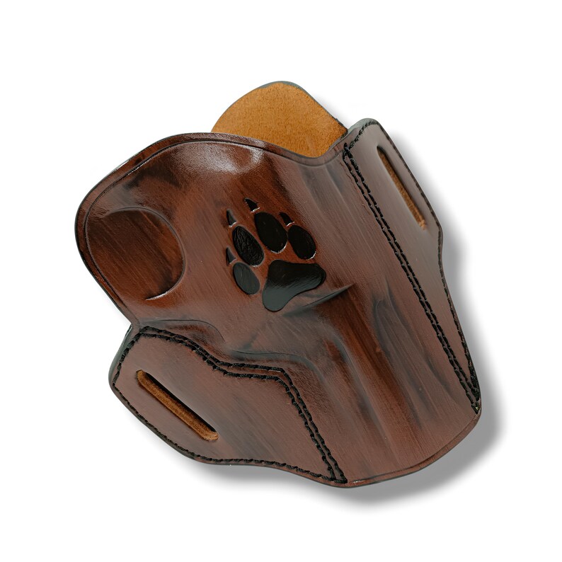 May include: A brown leather holster with a black paw print design. The holster has a glossy finish and black stitching around the edges. The interior is a lighter brown color.