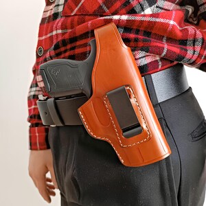 Op de afbeelding: Een bruinleren holster met witte stiksels, met een zwart pistool. De holster is bevestigd aan een zwarte riem. De persoon draagt een rood-zwart geruit overhemd en een zwarte broek.