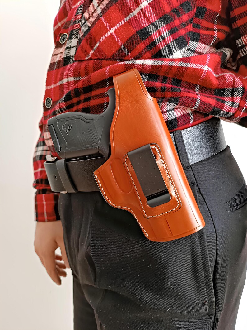 Op de afbeelding: Een bruinleren holster met witte stiksels, met een zwart pistool. De holster is bevestigd aan een zwarte riem. De persoon draagt een rood-zwart geruit overhemd en een zwarte broek.