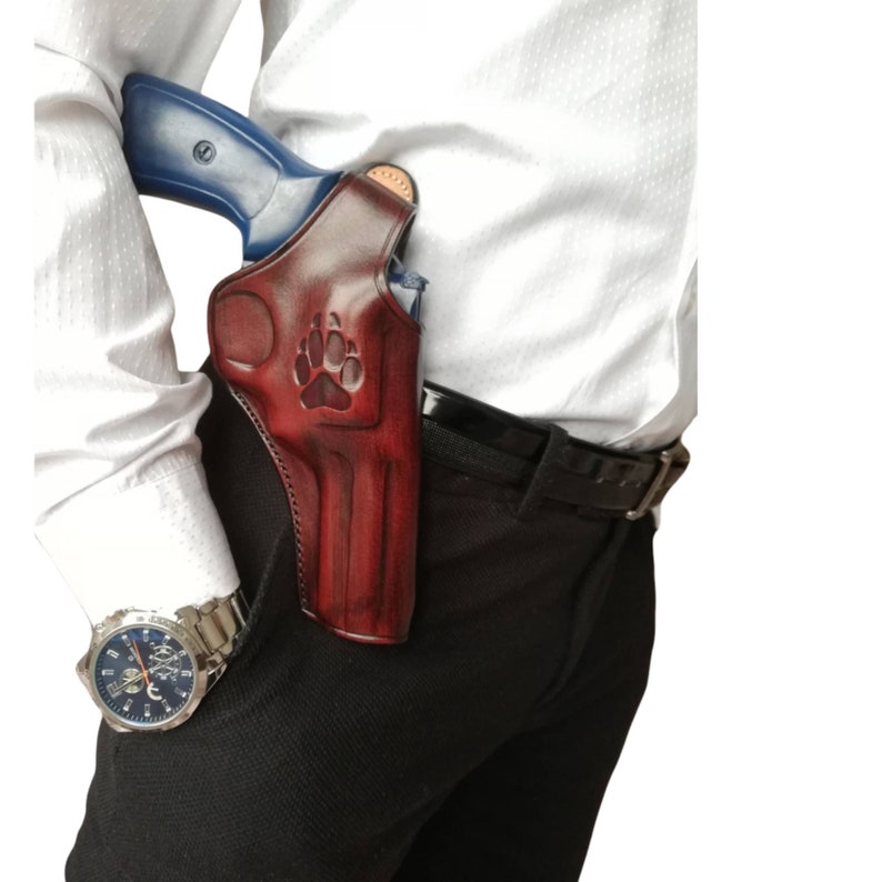 Peut inclure: Un &eacute;tui en cuir bordeaux avec un motif de patte, contenant un revolver bleu et argent&eacute;. L'&eacute;tui est attach&eacute; &agrave; une ceinture noire, port&eacute;e avec un pantalon noir et une chemise blanche. Une montre argent&eacute;e est visible au poignet.