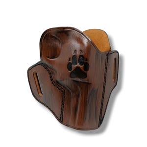 May include: A brown leather holster with a black paw print design. The holster has a textured finish and visible stitching. The interior is a lighter tan color. The holster is designed to hold a small object.