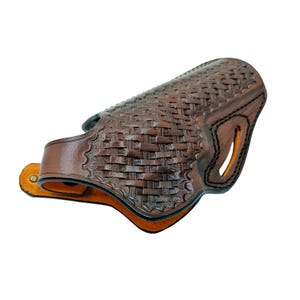 Pode incluir: Uma coldre de couro castanho com um padr&atilde;o de tecido de cesta. O coldre tem uma forma curva com uma presilha para cinto e um recorte para a pega. As bordas s&atilde;o costuradas com linha preta.