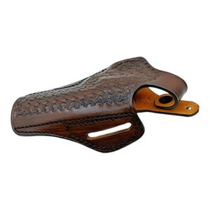 Op de afbeelding: Een bruin leren holster met een mandweefpatroon. De holster heeft een gebogen ontwerp met een riemlus en een drukknoopsluiting. Het leer heeft een rijke, donkerbruine kleur met een glanzende afwerking.