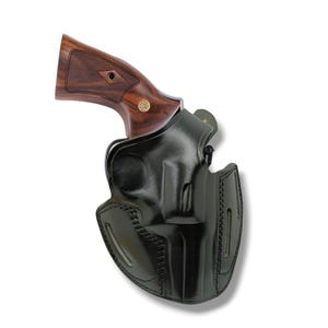 Op de afbeelding: Een zwart leren holster met een revolver. De revolver heeft een houten handvat met een bruine afwerking en een gouden embleem. Het holster is ontworpen om het vuurwapen veilig vast te houden.