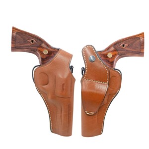 Può includere: Due revolver con impugnature in legno e fondine in pelle marrone. Le fondine sono cucite con filo bianco e hanno una chiusura a scatto. I revolver sono su sfondo bianco.