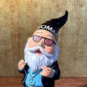 Peut inclure: Un gnome de jardin portant une veste en cuir noire, un jean bleu et des lunettes de soleil. Le gnome a une barbe blanche et un collier avec un symbole de paix. Le chapeau du gnome a le mot "OMAS" &eacute;crit dessus.