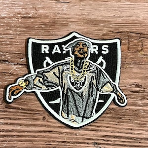 Könnte beinhalten: Ein gestickter Patch in Schwarzweiß, der einen Rapper zeigt, der ein Raiders-Trikot und eine goldene Kette trägt. Der Patch hat den Schriftzug "RAIDERS" in weißer Schrift auf einem schwarzen Schild.