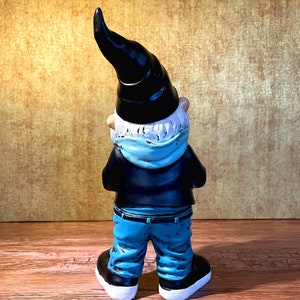 Peut inclure: Une figurine de gnome de jardin portant un chapeau noir, une &eacute;charpe bleue et un jean bleu. Le gnome est tourn&eacute; dos &agrave; la cam&eacute;ra et a une barbe blanche.