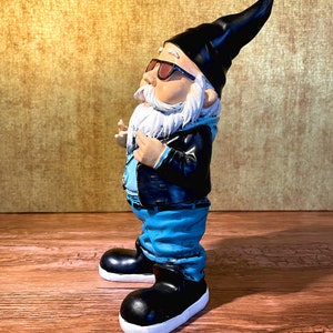 Peut inclure: Une figurine de gnome de jardin portant des lunettes de soleil, une veste en cuir noire, un jean bleu et des bottes noires. Le gnome a une barbe blanche et un chapeau pointu noir.