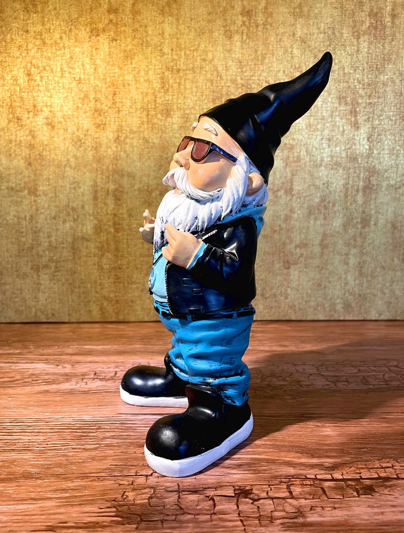 Peut inclure: Une figurine de gnome de jardin portant des lunettes de soleil, une veste en cuir noire, un jean bleu et des bottes noires. Le gnome a une barbe blanche et un chapeau pointu noir.